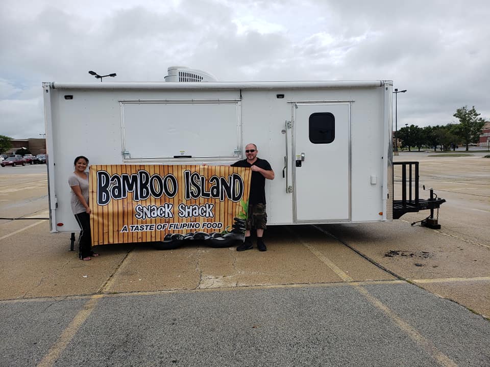 A man and a woman stand in front of a white food trailer, holding a large banner for "Bamboo Island Snack Shack: A Taste of Filipino Food."