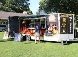 A black food truck with flame graphics and "Que It Up BBQ" branding, with a man standing at the service window and trees in the background.