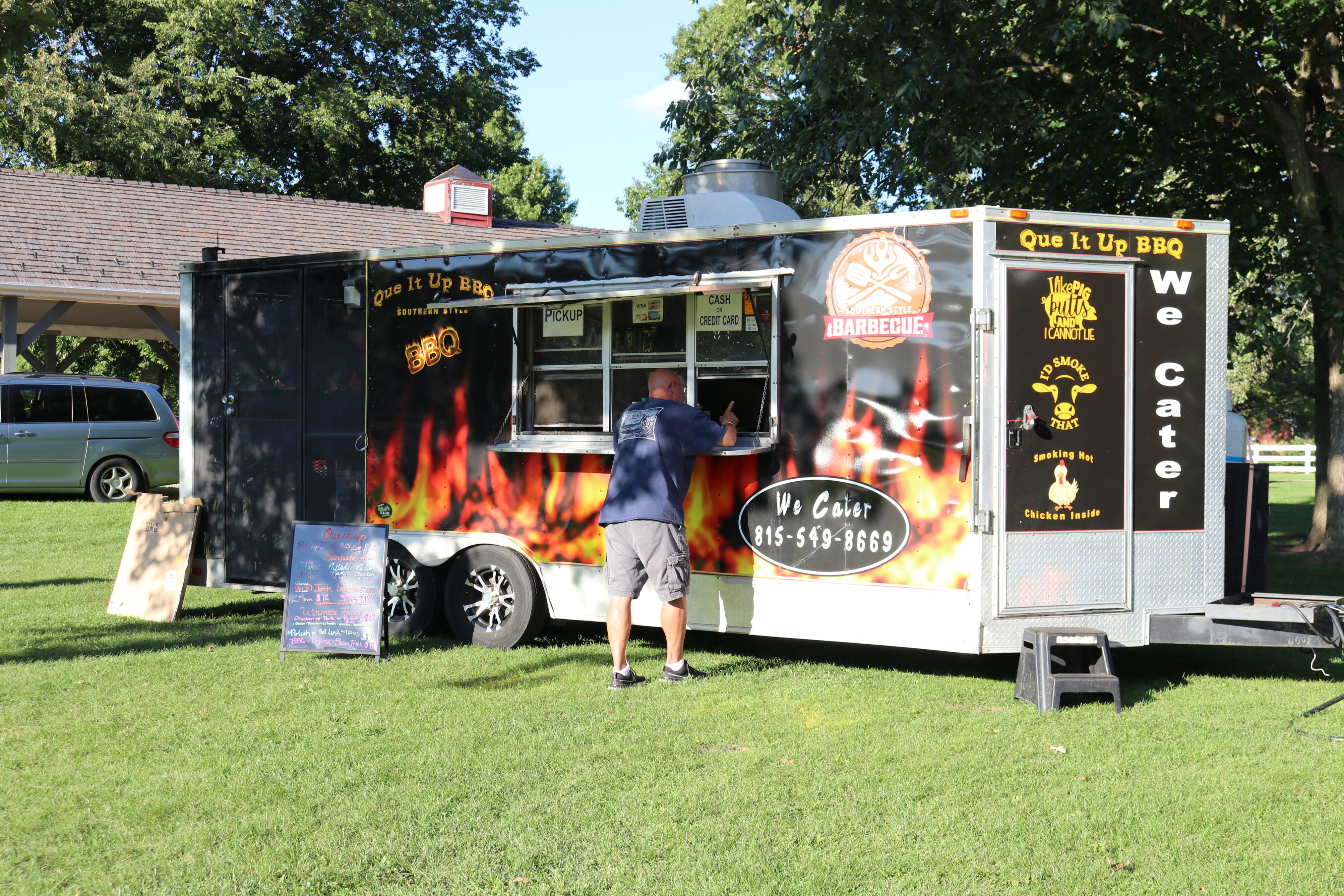 A black food truck with flame graphics and "Que It Up BBQ" branding, with a man standing at the service window and trees in the background.