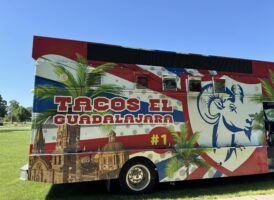 Side view of a "Tacos El Guadalajara #1" food truck, brightly decorated with red, white, and blue, palm trees, and a cityscape.