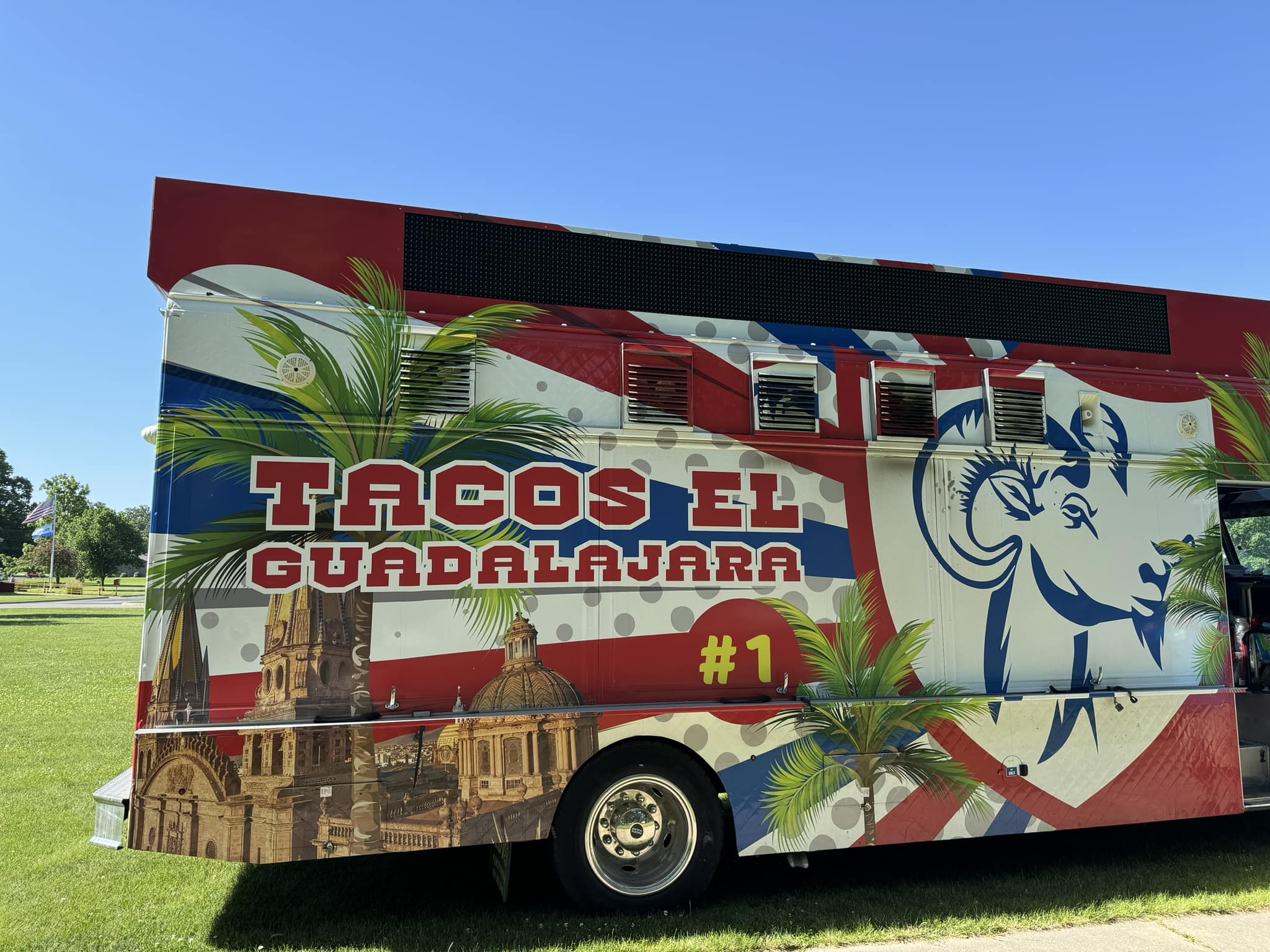Side view of a "Tacos El Guadalajara #1" food truck, brightly decorated with red, white, and blue, palm trees, and a cityscape.
