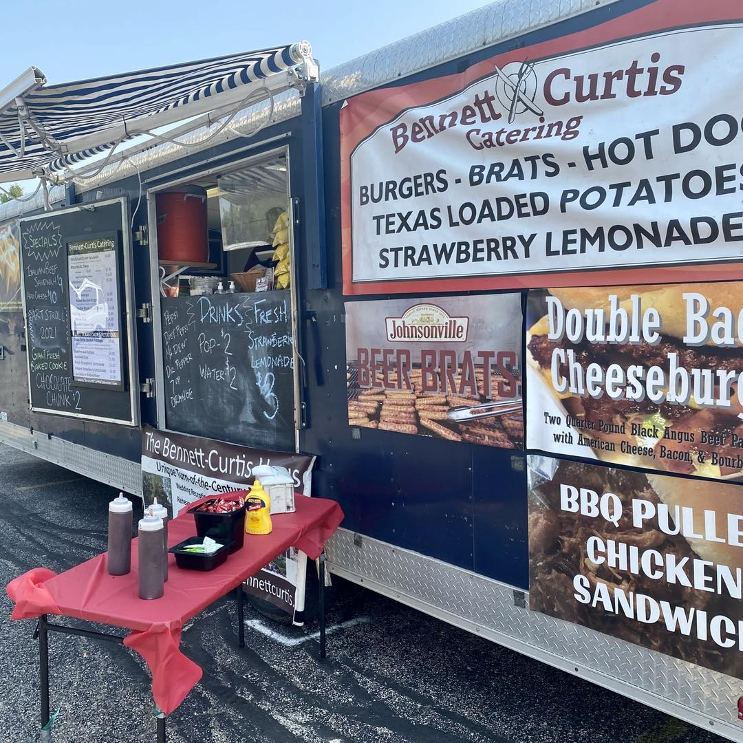 A "Bennett-Curtis Catering" food truck with its menu visible and a small table set up for condiments.