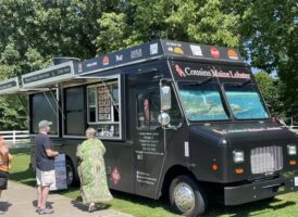 A black "Cousins Maine Lobster" food truck with its serving window open and several customers standing by.