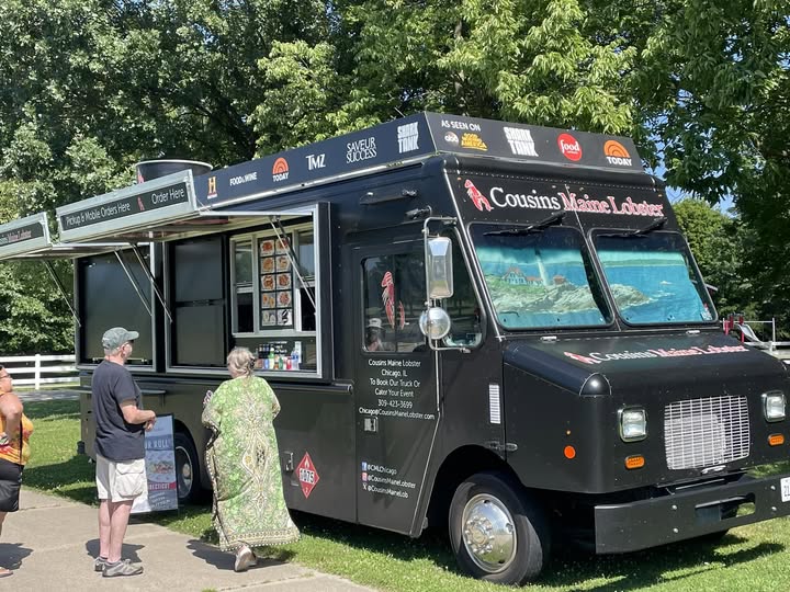 A black "Cousins Maine Lobster" food truck with its serving window open and several customers standing by.
