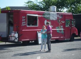 A red food truck named "Pasta Baby!" with two people standing in front of it.
