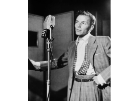 Black and white photo of Frank Sinatra in a suit, standing in front of a microphone in a recording studio.