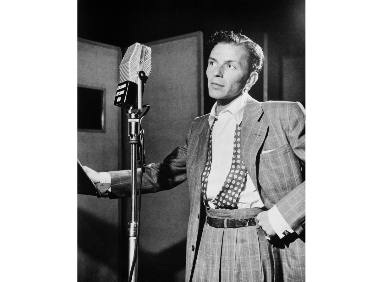 Black and white photo of Frank Sinatra in a suit, standing in front of a microphone in a recording studio.