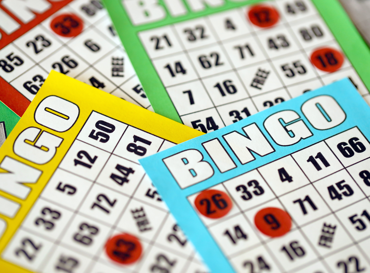 A stack of colorful Bingo cards laid out, with the top card in blue, showing the word "BINGO" across the top. Red plastic markers cover several numbers on the cards, indicating called numbers. Another yellow card is partially visible underneath, and a green and red card are further back.