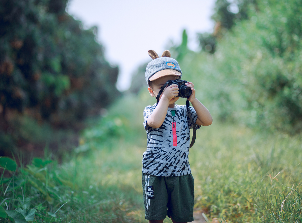 A small child, facing forward, stands in a grassy area holding a black camera up to their face as if taking a picture. The child is wearing a grey and tan baseball cap with two brown animal ears on top, a grey patterned t-shirt, and dark green shorts. The background is a soft blur of green foliage.