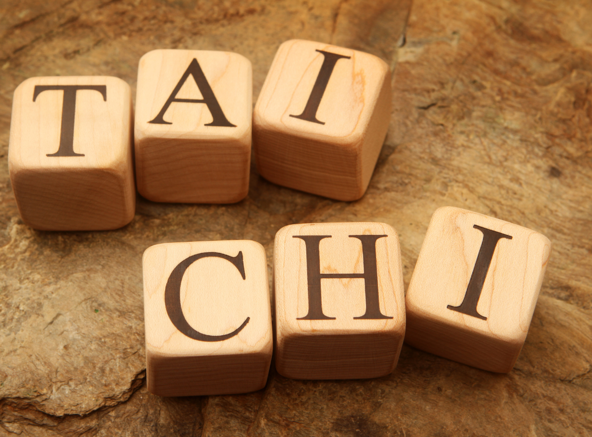 Six light-colored wooden alphabet blocks with dark uppercase letters. The blocks are arranged to spell "TAI" in the top row and "CHI" directly below it in the bottom row. They are resting on a rough, textured brown wooden surface, possibly tree bark or an old wooden board.