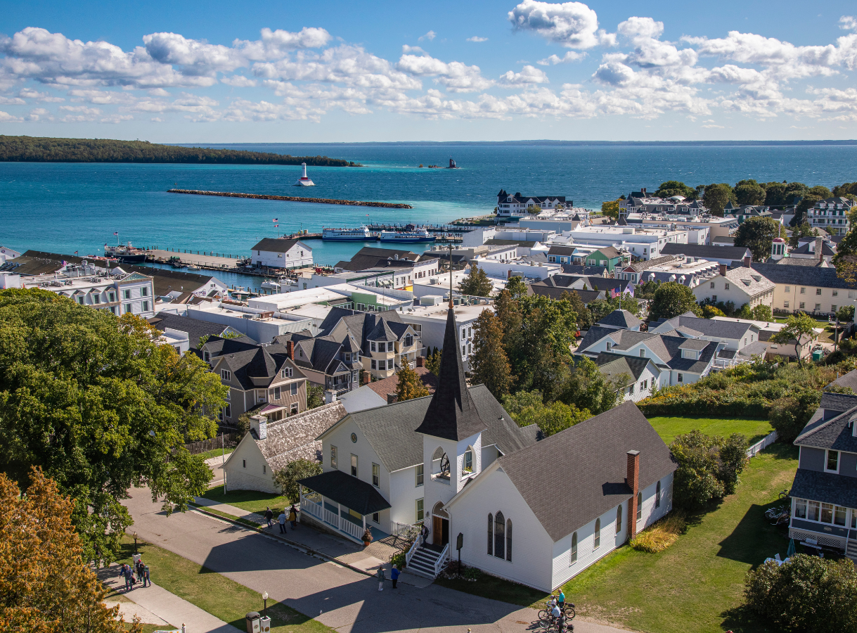An aerial or high-angle shot of a coastal town. In the foreground is a white church with a dark steeple and roof, surrounded by green trees and some houses. Beyond the town's buildings, a busy harbor is visible with ferries and boats docked. The turquoise-blue water of a large lake or strait stretches into the distance under a blue sky with white clouds, with land visible on the far horizon.