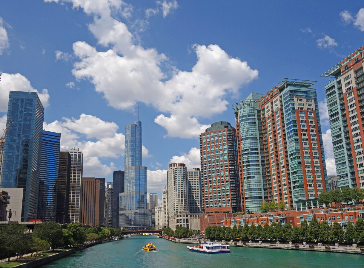 A wide-angle view of the Chicago River, which is a vibrant teal color, with various boats on its surface. The river is bordered on both sides by a dense skyline of tall buildings, ranging in color and architectural style, including the prominent blue-glass Trump International Hotel & Tower. A bright blue sky with numerous fluffy white clouds is above.