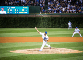 A baseball pitcher in a white and blue uniform throws a pitch from the mound during a game, with fans in the stands.