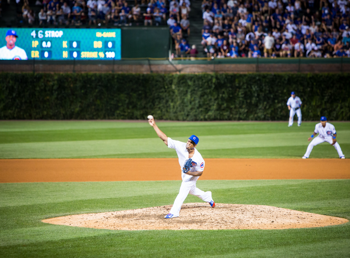 A baseball pitcher in a white and blue uniform throws a pitch from the mound during a game, with fans in the stands.