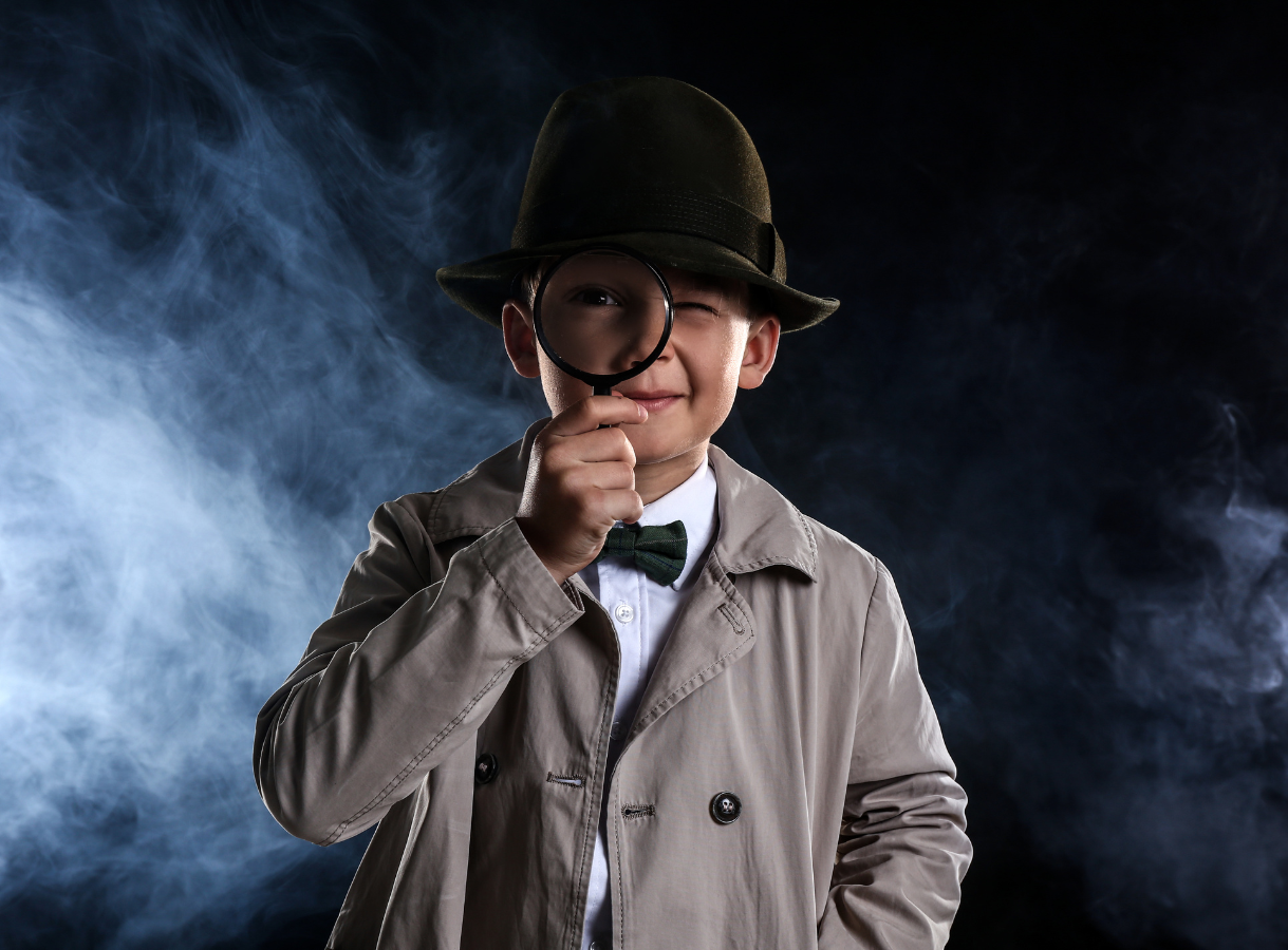 A child dressed as a detective, wearing a beige trench coat, a white shirt with a green bow tie, and a dark fedora hat. The child holds a magnifying glass up to their left eye, which is wide open, while their right eye is playfully winked shut. A smoky or misty dark background surrounds the child.