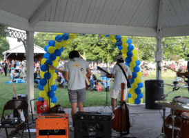 A view from behind two male musicians on a stage under a white gazebo. One plays an electric guitar, and the other an acoustic guitar. Behind them is a large arch made of alternating yellow and blue balloons. A crowd of people is seated on a grassy lawn in the background, enjoying the performance. Musical equipment, including amplifiers and a drum set, are visible in the foreground.