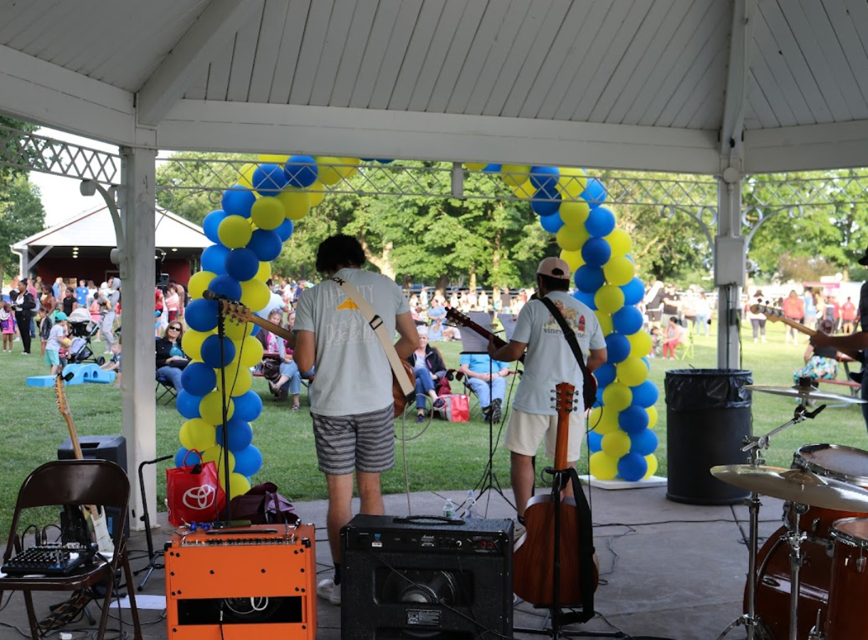 A view from behind two male musicians on a stage under a white gazebo. One plays an electric guitar, and the other an acoustic guitar. Behind them is a large arch made of alternating yellow and blue balloons. A crowd of people is seated on a grassy lawn in the background, enjoying the performance. Musical equipment, including amplifiers and a drum set, are visible in the foreground.