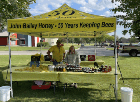 A yellow tent with "John Bailey Honey - 50 Years Keeping Bees" printed on it, under which a man and a woman stand behind tables laden with honey products and beeswax items.