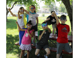 Six diverse children, dressed in superhero capes and masks, striking poses on and around a picnic table outdoors.