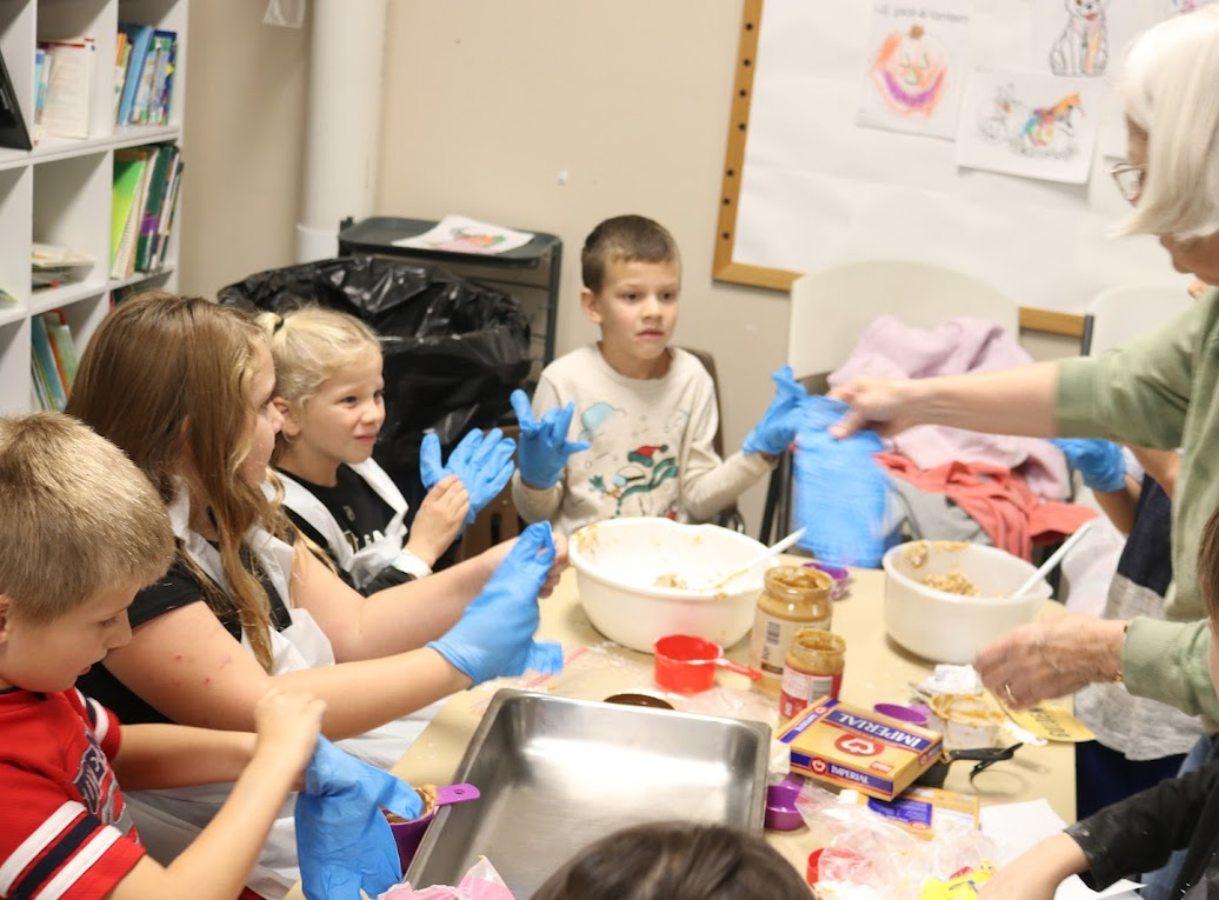Several children and an adult wearing blue gloves and aprons are gathered around a table with bowls and ingredients, participating in a cooking activity.