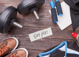 A flat lay shows two black dumbbells, a blue jump rope, a white notebook, brown sneakers, a black athletic shirt, and a red water bottle on a dark wooden background. A note in the center reads "Get fit!"