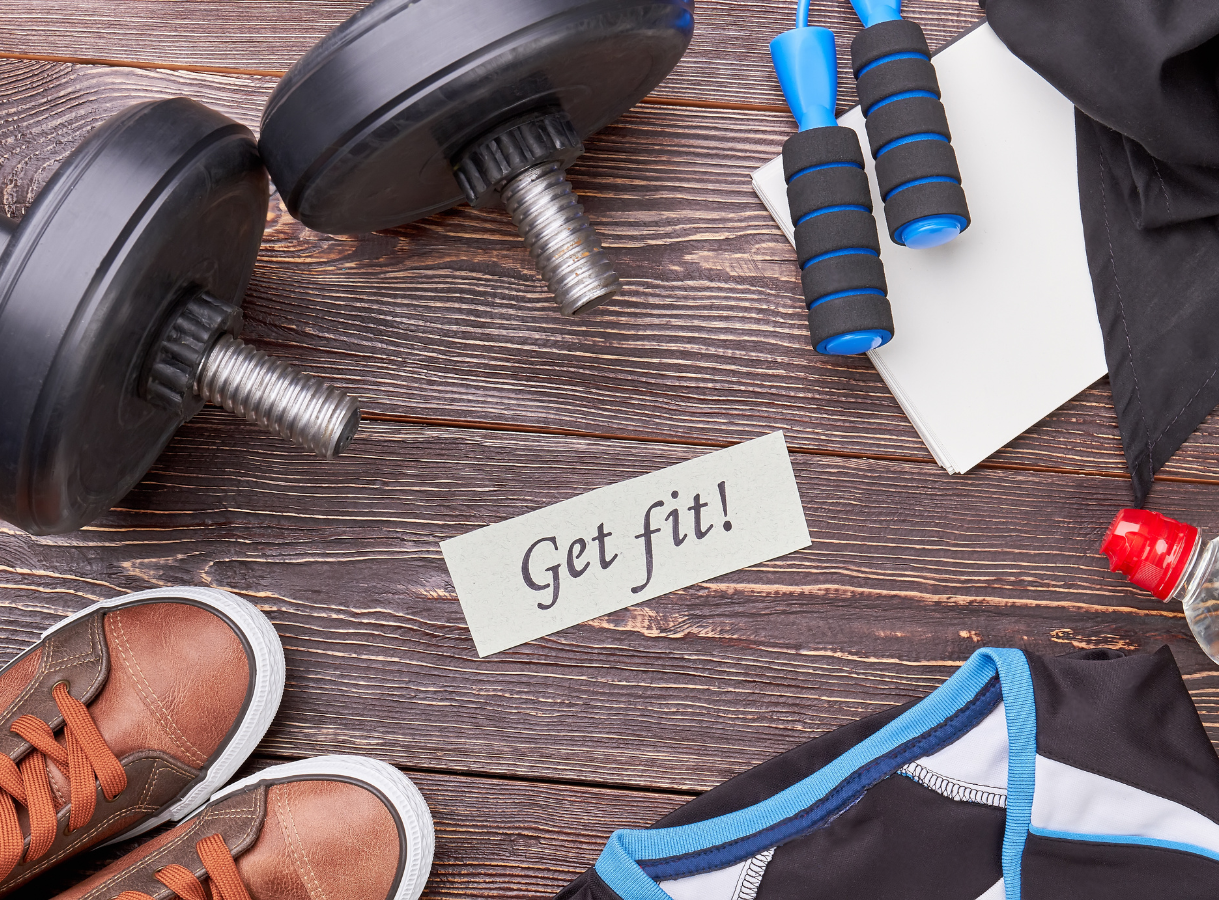 A flat lay shows two black dumbbells, a blue jump rope, a white notebook, brown sneakers, a black athletic shirt, and a red water bottle on a dark wooden background. A note in the center reads "Get fit!"