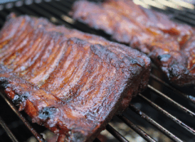 Two racks of sauced, reddish-brown BBQ ribs cooking on a black metal grill grate.