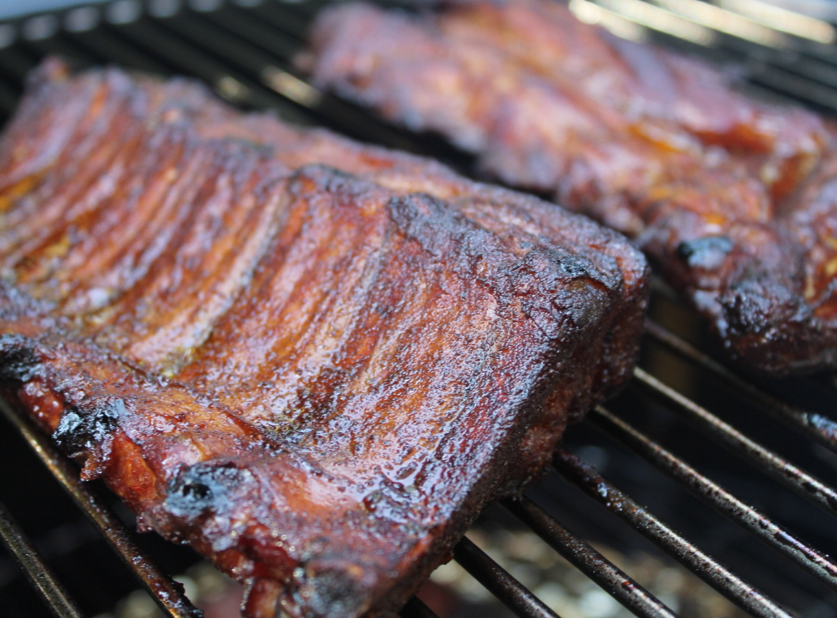 Two racks of sauced, reddish-brown BBQ ribs cooking on a black metal grill grate.