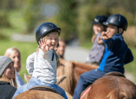 A young girl in a black helmet on a horse smiles and waves at the camera, with other children and adults also on horseback in the background.