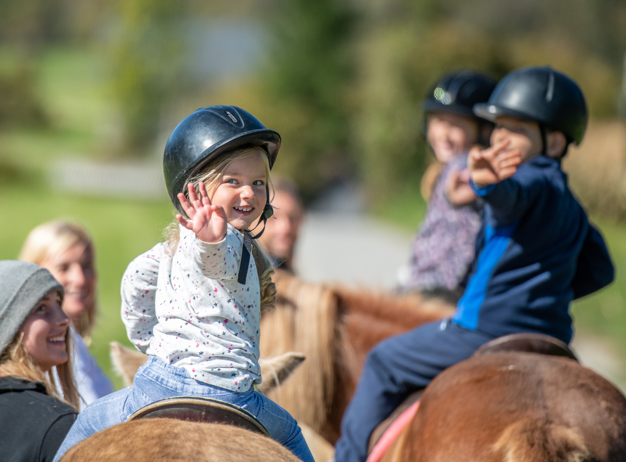 A young girl in a black helmet on a horse smiles and waves at the camera, with other children and adults also on horseback in the background.
