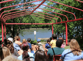 A group of people, including adults and children, sit in an open-air vehicle with a red frame, looking out at a wide body of water bordered by green trees.