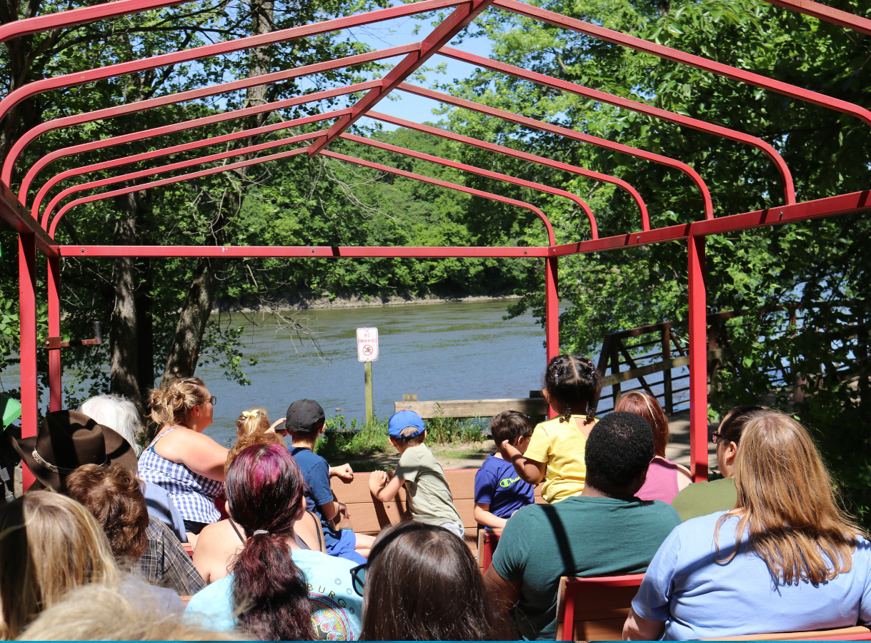 A group of people, including adults and children, sit in an open-air vehicle with a red frame, looking out at a wide body of water bordered by green trees.