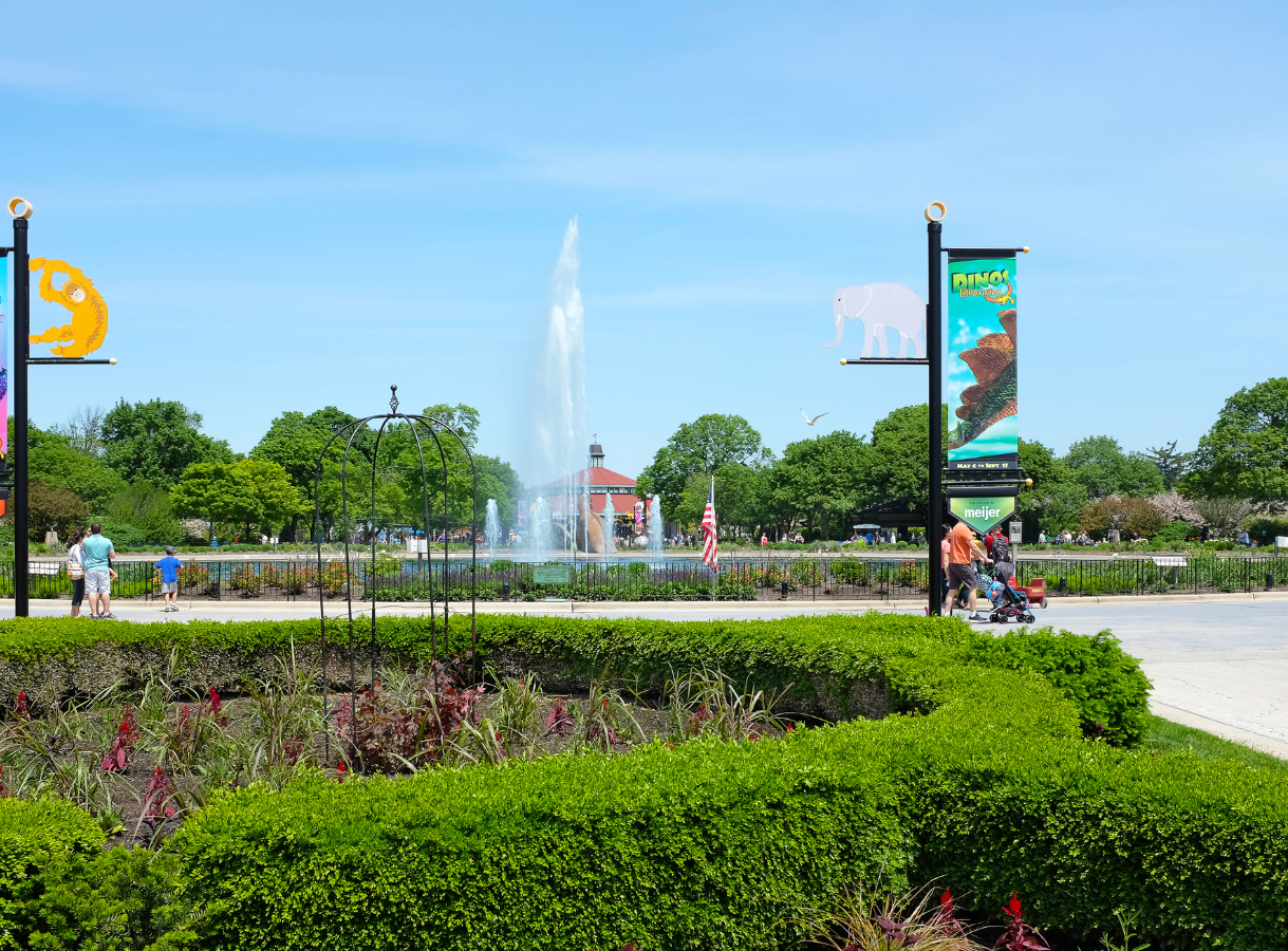 A wide shot of the Roosevelt Fountain at Brookfield Zoo, with a tall geyser of water spraying into the air. Lush green hedges and flowerbeds are in the foreground, and people can be seen walking on paved paths around the fountain. Colorful banners depicting animals are visible on lampposts.