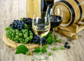 Two glasses of wine, one filled with white wine and one with red wine, stand on a light wooden surface. Behind them are two wine bottles and a small wooden barrel. To the left, a wooden cutting board holds large bunches of green and dark purple grapes. Some loose grapes and a vine leaf are scattered in the foreground.