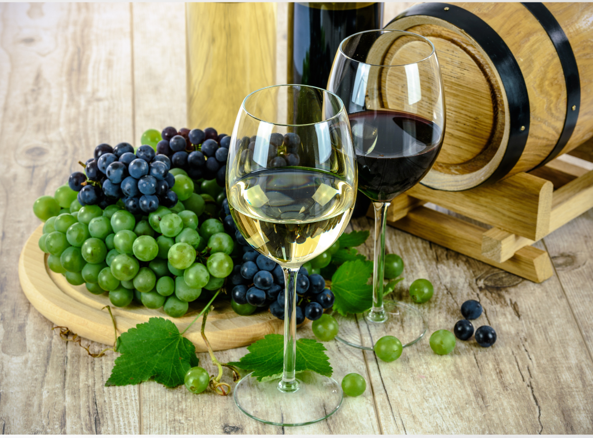 Two glasses of wine, one filled with white wine and one with red wine, stand on a light wooden surface. Behind them are two wine bottles and a small wooden barrel. To the left, a wooden cutting board holds large bunches of green and dark purple grapes. Some loose grapes and a vine leaf are scattered in the foreground.