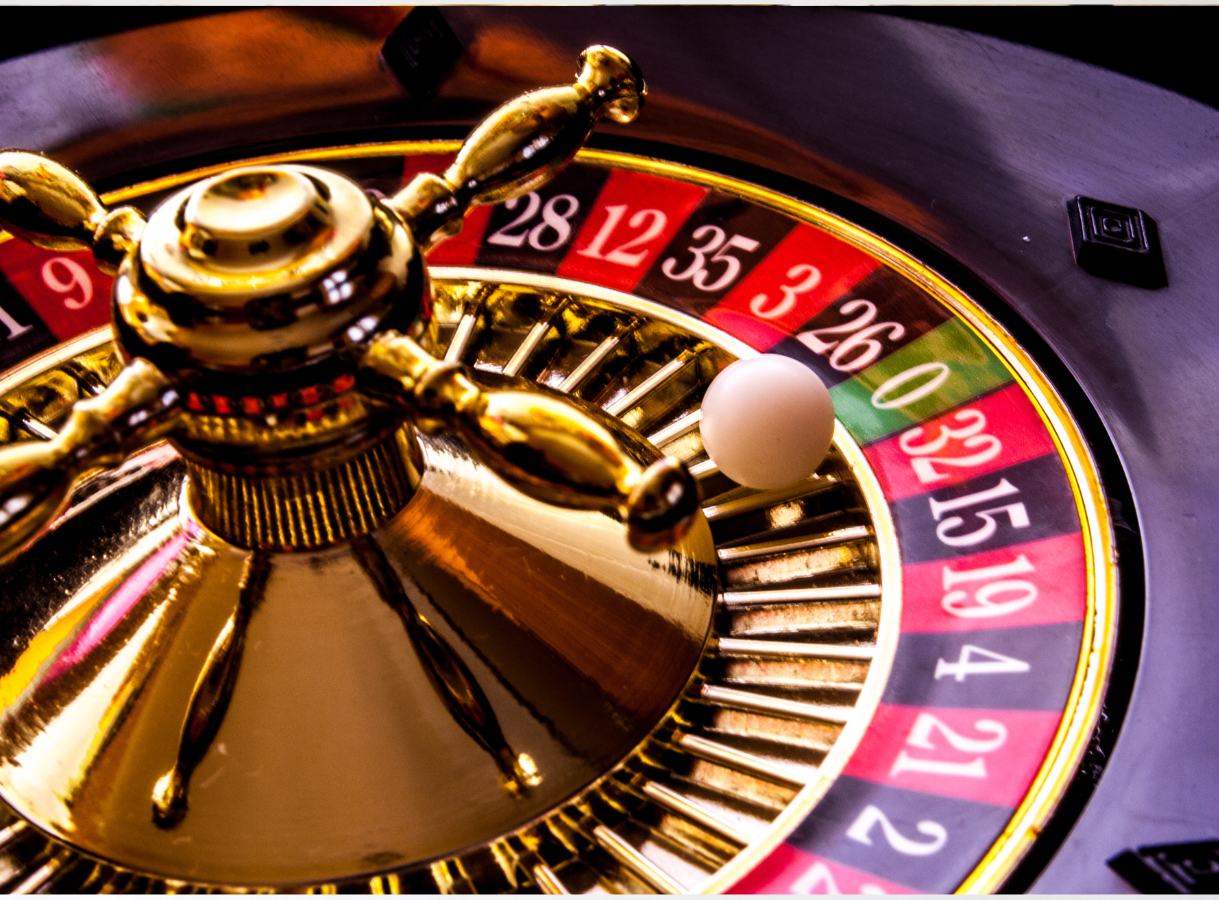 A close-up, high-angle view of a roulette wheel. The numbers on the wheel are visible, including 0 in green, and a series of red and black numbers. A small white ball is resting in the pocket for the number 0. The central golden spindle and its radiating spokes are prominent, reflecting light. The wheel's surface appears glossy and dark around the numbered pockets.