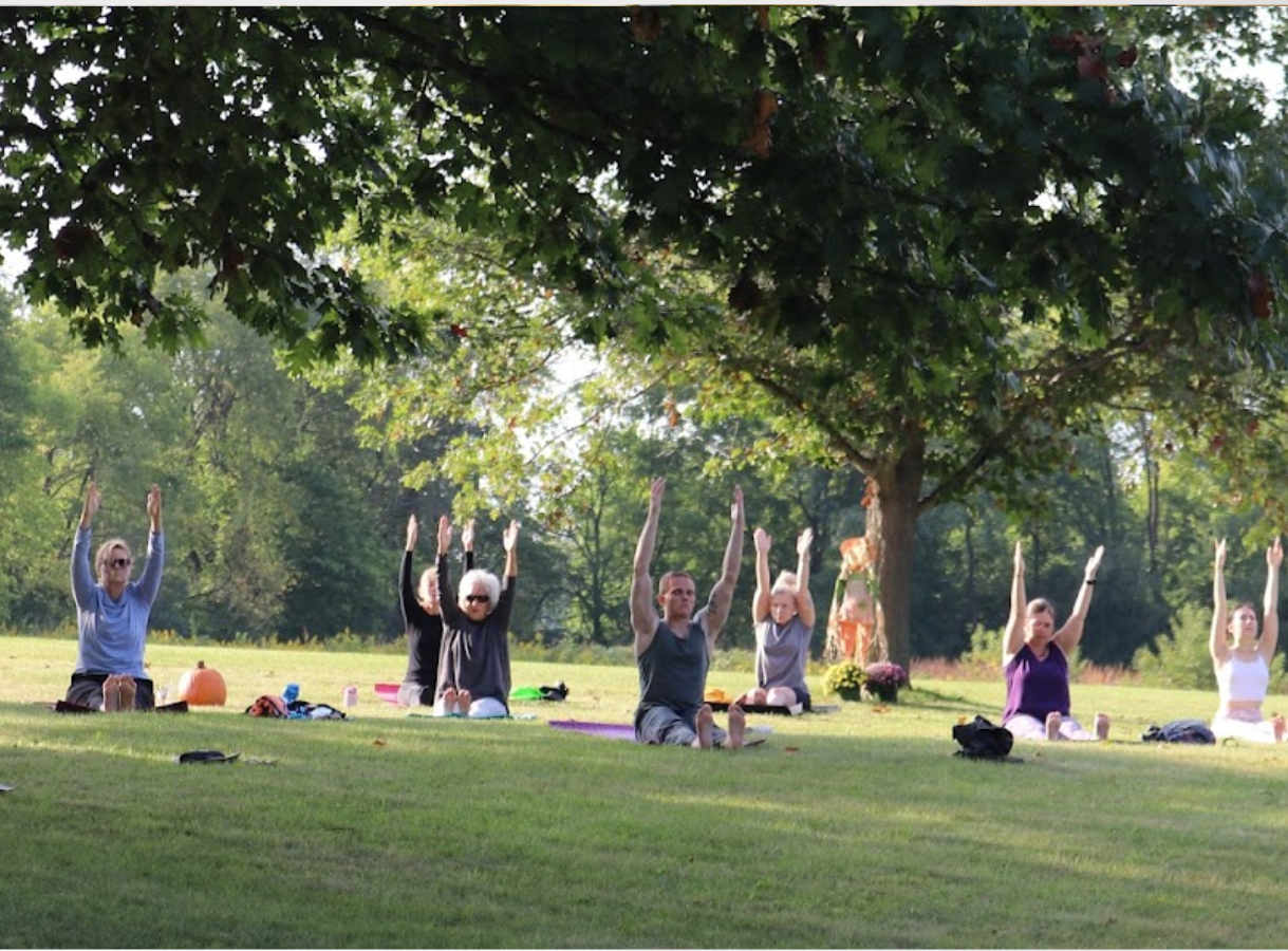 A group of approximately eight people are sitting on yoga mats on a grassy lawn with their arms extended upwards in a yoga pose. They are spaced out under the shade of a large tree with lush green foliage. In the background, more trees and greenery are visible, creating a peaceful outdoor setting. Some personal items like water bottles and a pumpkin are scattered near the participants.