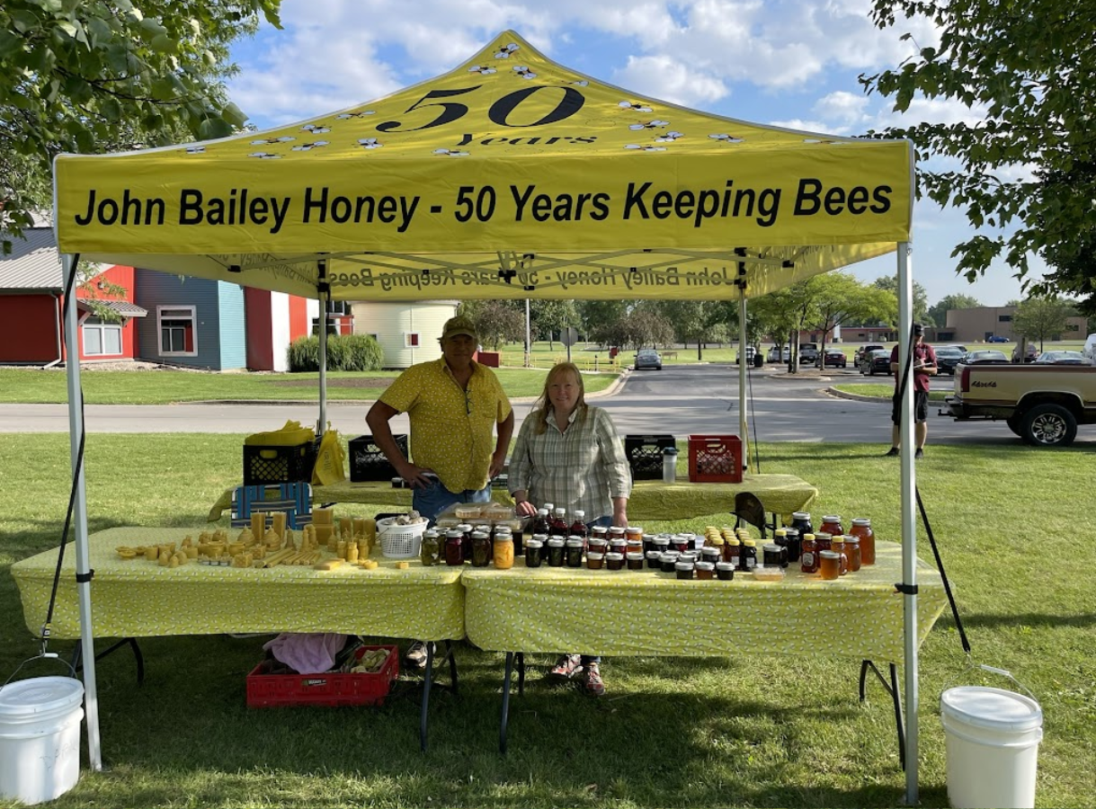 A yellow tent with "John Bailey Honey - 50 Years Keeping Bees" printed on it, under which a man and a woman stand behind tables laden with honey products and beeswax items.