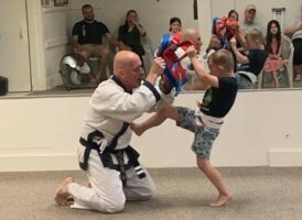 A young boy in a blue t-shirt and camouflage shorts practices a kick on a man in a white taekwondo uniform holding a kicking shield, with spectators in the background.