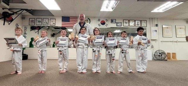A group of young martial arts students in white gis holding certificates and broken boards, standing with an instructor.