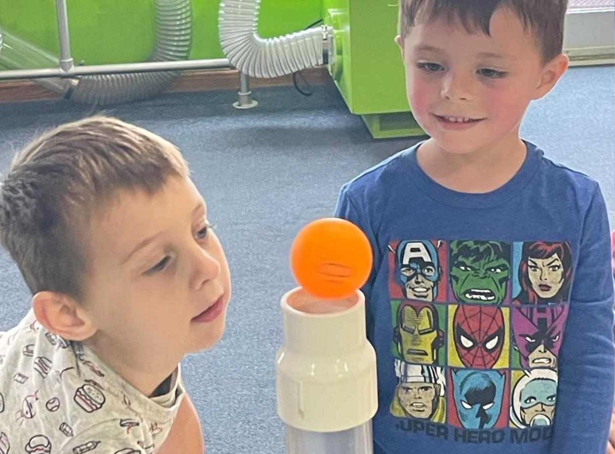 Two young boys look intently at an orange ball levitating above a white tube, likely at an interactive science exhibit.